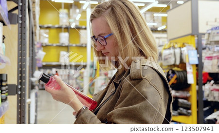 Blonde woman wearing glasses and leather jacket choosing cosmetics in supermarket reading label on bottle, shelves with various products in background Blonde woman wearing glasses and leather jacket choosing cosmetics in supermarket reading label on bottle, shelves with various products in background 123507941