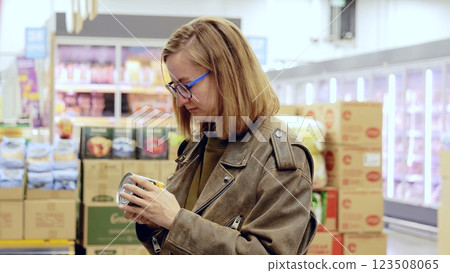 Young woman wearing glasses and leather jacket choosing canned food, carefully reading product information on label in supermarket aisle 123508065