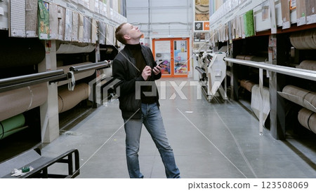 Customer uses a smartphone to compare carpets while shopping for floor coverings in a hardware store, planning home improvement. A man in a hardware store choosing flooring. Customer uses a smartphone to compare carpets while shopping for floor coverings in a hardware store, planning home improvement. A man in a hardware store choosing flooring. 123508069