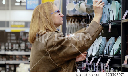 Blonde woman wearing glasses choosing plates in a houseware store, reaching for a light blue plate on a shelf, shopping for kitchenware in a retail environment Blonde woman wearing glasses choosing plates in a houseware store, reaching for a light blue plate on a shelf, shopping for kitchenware in a retail environment 123508071