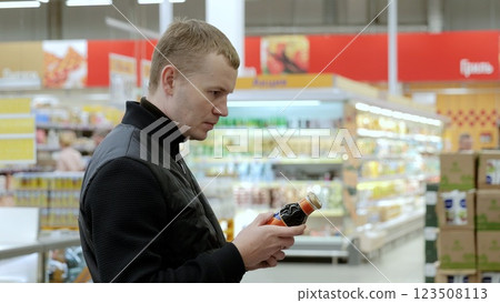 Shopper reads the label on a bottle while comparing beverages in the supermarket aisle. A customer in black studies the label on a bottle of sauce 123508113