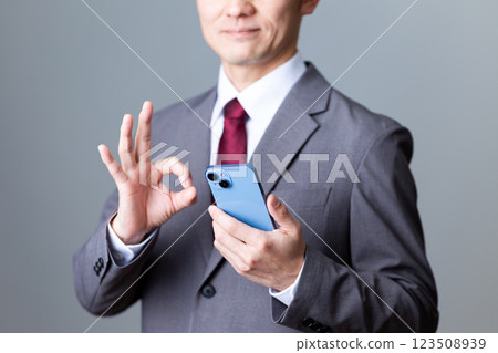 一位中年男性商人身穿灰色西裝，手拿智慧型手機，做著圓形手勢 123508939