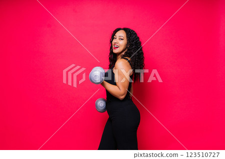 Smiling african american female doing hand exercises with dumbbells on red studio background. Smiling african american female doing hand exercises with dumbbells on red studio background. 123510727