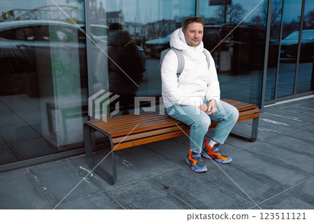 Smiling young white man in casual cothes using mobile phone outdoors in the city, sitting on a bench 123511121