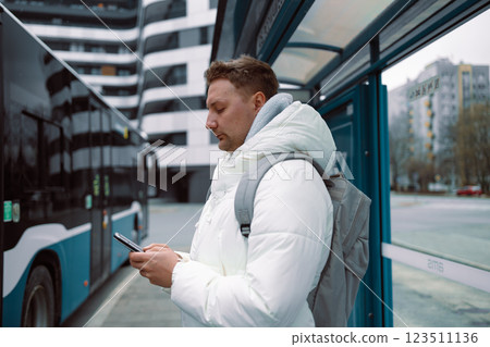 Young stylish man with smart phone on bus stop and typing sms stand at public transport bus station taking bus with headphones and mobile smart phone in winter or autumn day with backpack Young stylish man with smart phone on bus stop and typing sms stand at public transport bus station taking bus with headphones and mobile smart phone in winter or autumn day with backpack 123511136