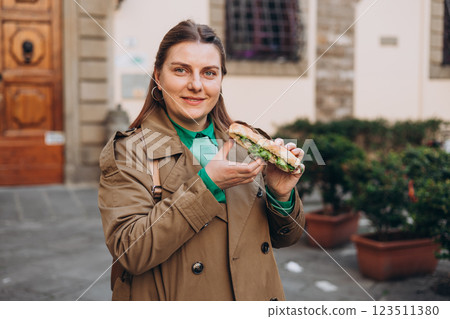 Young happy 30s woman eating sandwich on the city street. Urban food festival. Beautiful hungry girl eating tasty burger outdoor Young happy 30s woman eating sandwich on the city street. Urban food festival. Beautiful hungry girl eating tasty burger outdoor 123511380