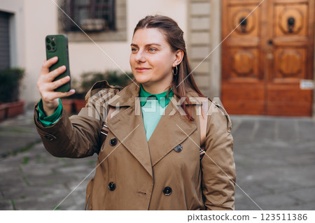Picture of pretty young woman holding phone in hands on the street. Redhead girl making selfie photo on urban background. Lifestyle, travel, tourism, technology, blogging, communication concept. Picture of pretty young woman holding phone in hands on the street. Redhead girl making selfie photo on urban background. Lifestyle, travel, tourism, technology, blogging, communication concept. 123511386