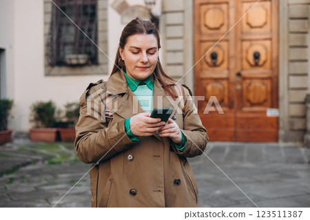 Happy cheerful young woman with backpack walking on city street checks her smartphone. Portrait of beautiful 30s girl using smartphone outdoors. Urban lifestyle concept. Happy cheerful young woman with backpack walking on city street checks her smartphone. Portrait of beautiful 30s girl using smartphone outdoors. Urban lifestyle concept. 123511387