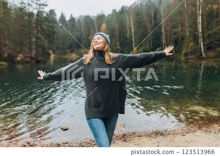 Happy woman raising hands up on the banks of the lake. Young 30s woman hiker in front smiling happy, hiking in woods, warm autumn day. 123513966