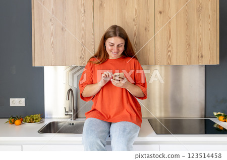 Portrait of smiling housewife is sitting on desk and using phone in modern kitchen. Home, people concept. Copy space. 30s Woman watching recipe on social media, video tutorial how to prepare meal. 123514458