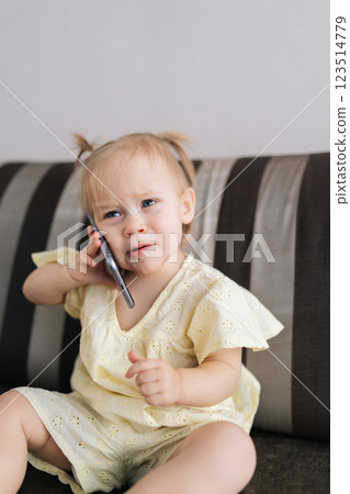 Vertical portrait of serious funny toddler girl listening intently to smartphone, pretending to make call while sitting on couch. Cheerful child expressing playful communication skills at home. 123514779
