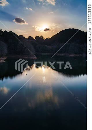 The temperature is rising and it's starting to feel like spring at Nara Nunome Dam. The sun sets over the mountains on the opposite shore, painting the sky and lake surface golden. 123515316