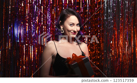 Holiday party. Surprise gift. Birthday greeting idea. Special occasion. Excited pretty woman fancy dress holding present box on red shimmering cascade curtain background. Holiday party. Surprise gift. Birthday greeting idea. Special occasion. Excited pretty woman fancy dress holding present box on red shimmering cascade curtain background. 123517359
