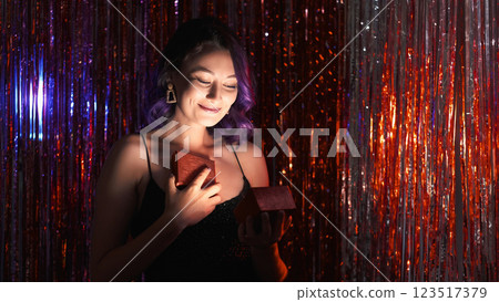 Precious gift. Holiday celebration. Special event. Wonderful party night. Delighted happy woman fancy dress open present box on red shimmering cascade curtain background. Precious gift. Holiday celebration. Special event. Wonderful party night. Delighted happy woman fancy dress open present box on red shimmering cascade curtain background. 123517379