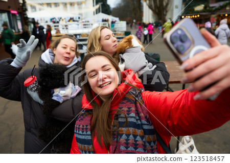 Friends take a joyful selfie with pets at a winter festival in a lively outdoor setting Friends take a joyful selfie with pets at a winter festival in a lively outdoor setting 123518457