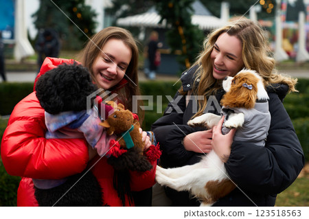 Two young women enjoy winter festivities while holding their adorable dogs in a park decorated for the holidays Two young women enjoy winter festivities while holding their adorable dogs in a park decorated for the holidays 123518563
