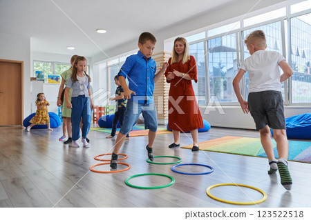 Small nursery school children with female teacher on floor indoors in classroom, doing exercise. Jumping over hula hoop circles track on the floor. Small nursery school children with female teacher on floor indoors in classroom, doing exercise. Jumping over hula hoop circles track on the floor. 123522518