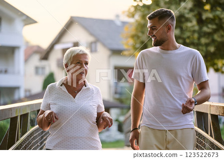 A handsome man and an older woman share a serene walk in nature, crossing a beautiful bridge against the backdrop of a stunning sunset, embodying the concept of a healthy and vibrant intergenerational 123522673