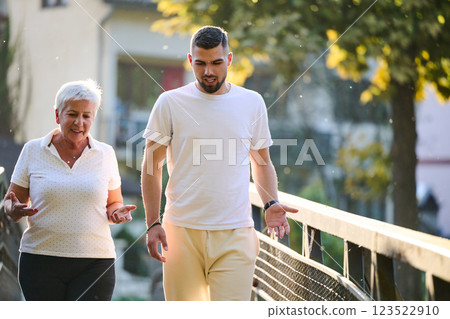 A handsome man and an older woman share a serene walk in nature, crossing a beautiful bridge against the backdrop of a stunning sunset, embodying the concept of a healthy and vibrant intergenerational 123522910