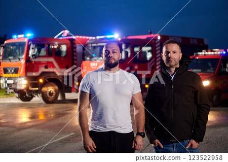 Group of firefighters, dressed in civilian clothing, stand in front of fire trucks during the night, showcasing a moment of camaraderie and unity among the team as they reflect on their duties and the Group of firefighters, dressed in civilian clothing, stand in front of fire trucks during the night, showcasing a moment of camaraderie and unity among the team as they reflect on their duties and the 123522958
