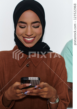 An elegant Arab woman, adorned in a hijab, engages with modernity as she uses a smartphone, the juxtaposition of traditional attire against contemporary technology captured in the isolated setting 123522968