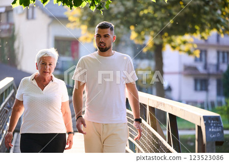 A handsome man and an older woman share a serene walk in nature, crossing a beautiful bridge against the backdrop of a stunning sunset, embodying the concept of a healthy and vibrant intergenerational 123523086