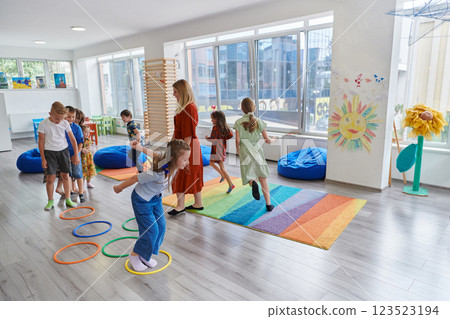 Small nursery school children with female teacher on floor indoors in classroom, doing exercise. Jumping over hula hoop circles track on the floor. 123523194