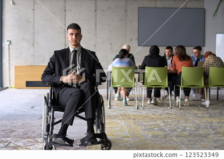 Business Director in a Suit Using a Wheelchair with Colleagues Conducting a Meeting Behind Him 123523349