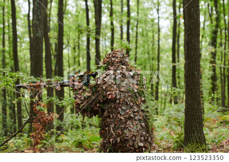 A highly skilled elite sniper, camouflaged in the dense forest, stealthily maneuvers through dangerous woodland terrain on a covert and precise mission 123523350