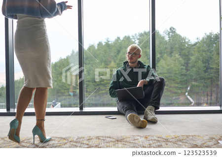 A blonde man sitting casually on the floor of a modern office, leaning against a window as he uses his laptop, with his attractive female colleague standing nearby A blonde man sitting casually on the floor of a modern office, leaning against a window as he uses his laptop, with his attractive female colleague standing nearby 123523539