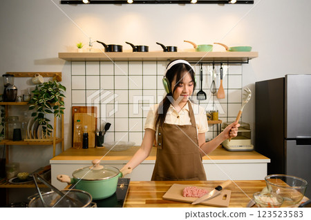 Cheerful asian woman cooking spaghetti in a cozy kitchen and listening to music with headphones 123523583