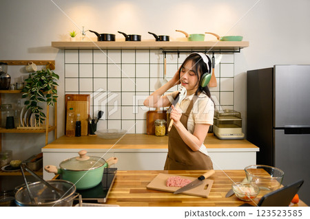 Cheerful asian woman cooking spaghetti in a cozy kitchen and listening to music with headphones 123523585