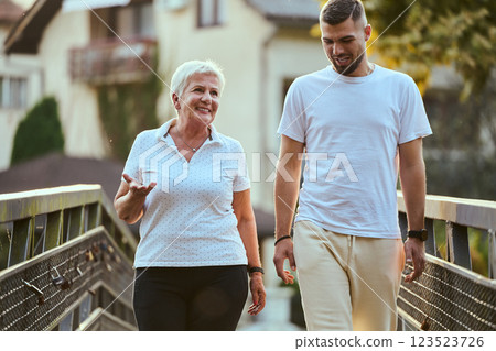 A handsome man and an older woman share a serene walk in nature, crossing a beautiful bridge against the backdrop of a stunning sunset, embodying the concept of a healthy and vibrant intergenerational 123523726