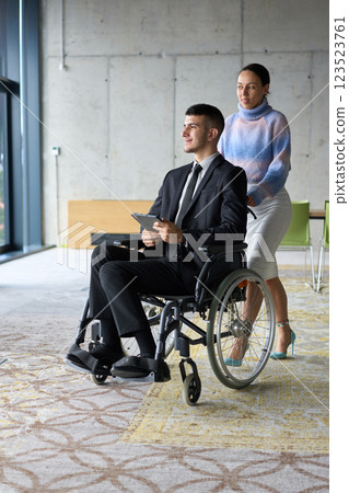 In a professional office setting, a businesswoman helping her director, who is sitting in a wheelchair and using a tablet, while their colleagues collaborate in the background. 123523761