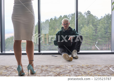 A blonde man sitting casually on the floor of a modern office, leaning against a window as he uses his laptop, with his attractive female colleague standing nearby 123523787