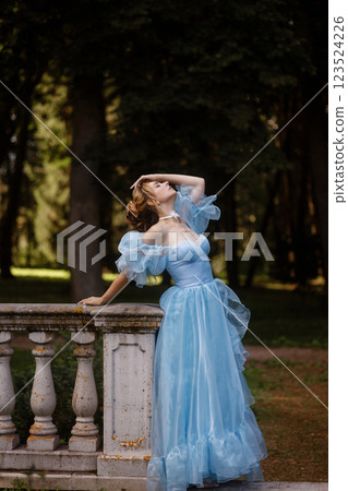 Romantic Woman in a Light Blue Gown Posing on a stone balustrade in a picturesque garden. 123524226