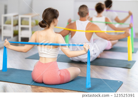 Back view of slim flexible athletic young woman sitting on sports mat practicing pilates during group exercises with elastic band in sports club 123525391