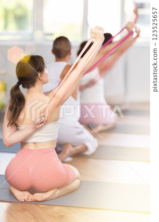 Rear view of young energetic athletic woman doing pilates kneeling on mat using resistance bands during group classes in fitness room 123525657