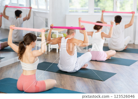 Back view of young girl with group of young adults maintaining physical health practicing pilates with resistance band sitting on gymnastics mat at fitness center 123526046