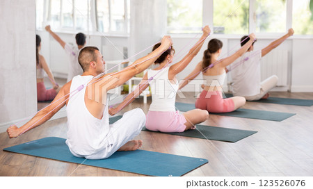 Young men and women doing shoulder workout with resistance band sitting cross-legged 123526076