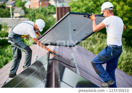 Workers building solar panel system on roof of house. Two men installers in helmets carrying photovoltaic solar module outdoors. Alternative, green and renewable energy generation concept. 123530818