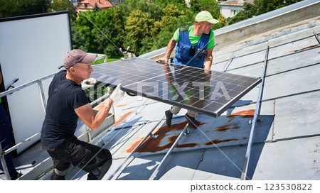 Workers building solar panel system on metal rooftop of house with assistance of crane lift. Two men installers carrying photovoltaic solar module outdoors. Renewable energy generation concept. 123530822