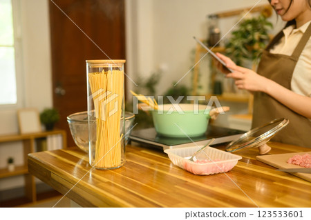 Dry spaghetti in a glass jar, meat, and boiling pot on kitchen counter with woman checking recipe on digital tablet 123533601