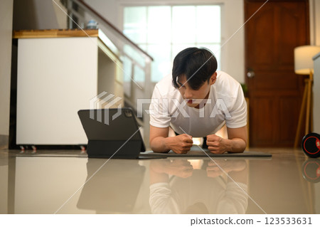 Focused young man doing plank exercise on a mat and watching training videos on digital tablet 123533631