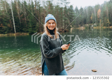 Phone Communication. Check social networks. 30s Traveler woman in hat standing with phone near the majestic lake. Travel, adventure. Concept of an active lifestyle. 123535302
