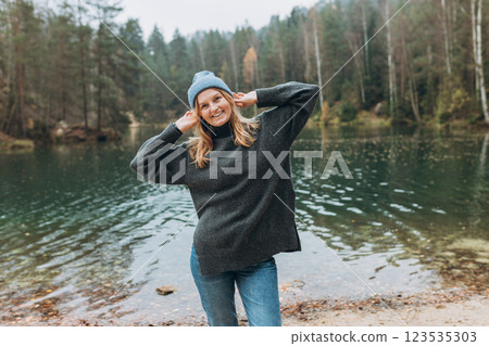 Happy woman in hat posing up on the banks of the lake. Young 30s woman hiker smiling, hiking in woods, warm autumn day. Travel, adventure, people banner. Concept of an active lifestyle 123535303