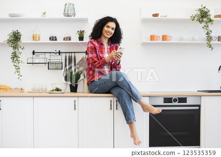 African American woman sitting on kitchen counter, wearing casual attire, using smartphone, smiling, modern kitchen decor. 123535359