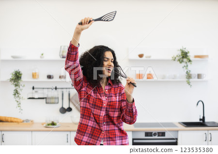 Smiling African woman in twenties holding kitchen tools, singing in modern bright kitchen setup. Expressing joy, creativity, and fun through music and dance. Smiling African woman in twenties holding kitchen tools, singing in modern bright kitchen setup. Expressing joy, creativity, and fun through music and dance. 123535380