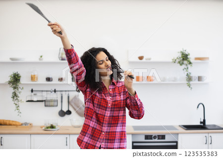 Smiling African woman in twenties holding kitchen tools, singing in modern bright kitchen setup. Expressing joy, creativity, and fun through music and dance. 123535383