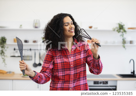 Young African woman, wearing vibrant plaid shirt, standing in kitchen, holding utensils, smiling energetically, expressing creativity and joy. 123535385